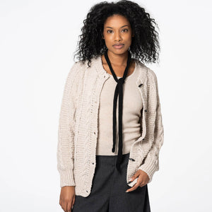 Woman wearing a beige cardigan over a white shirt with a black tie, standing against a white background.
