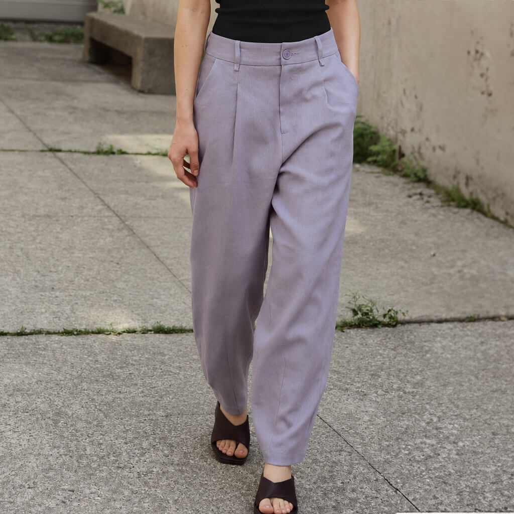 A person wearing loose, high-waisted light lavender pants with a black top and dark brown slide sandals, standing on a concrete sidewalk.