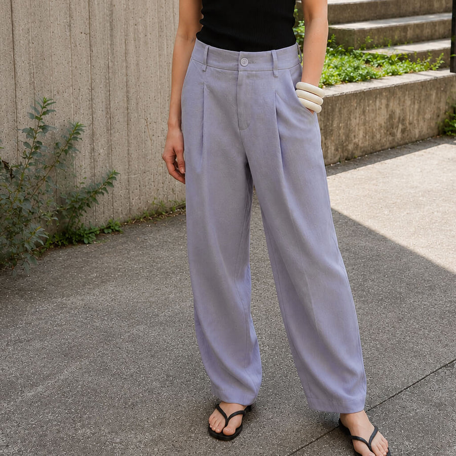 A person wearing loose, high-waisted light lavender pants with a black top and dark brown slide sandals, standing on a concrete sidewalk.
