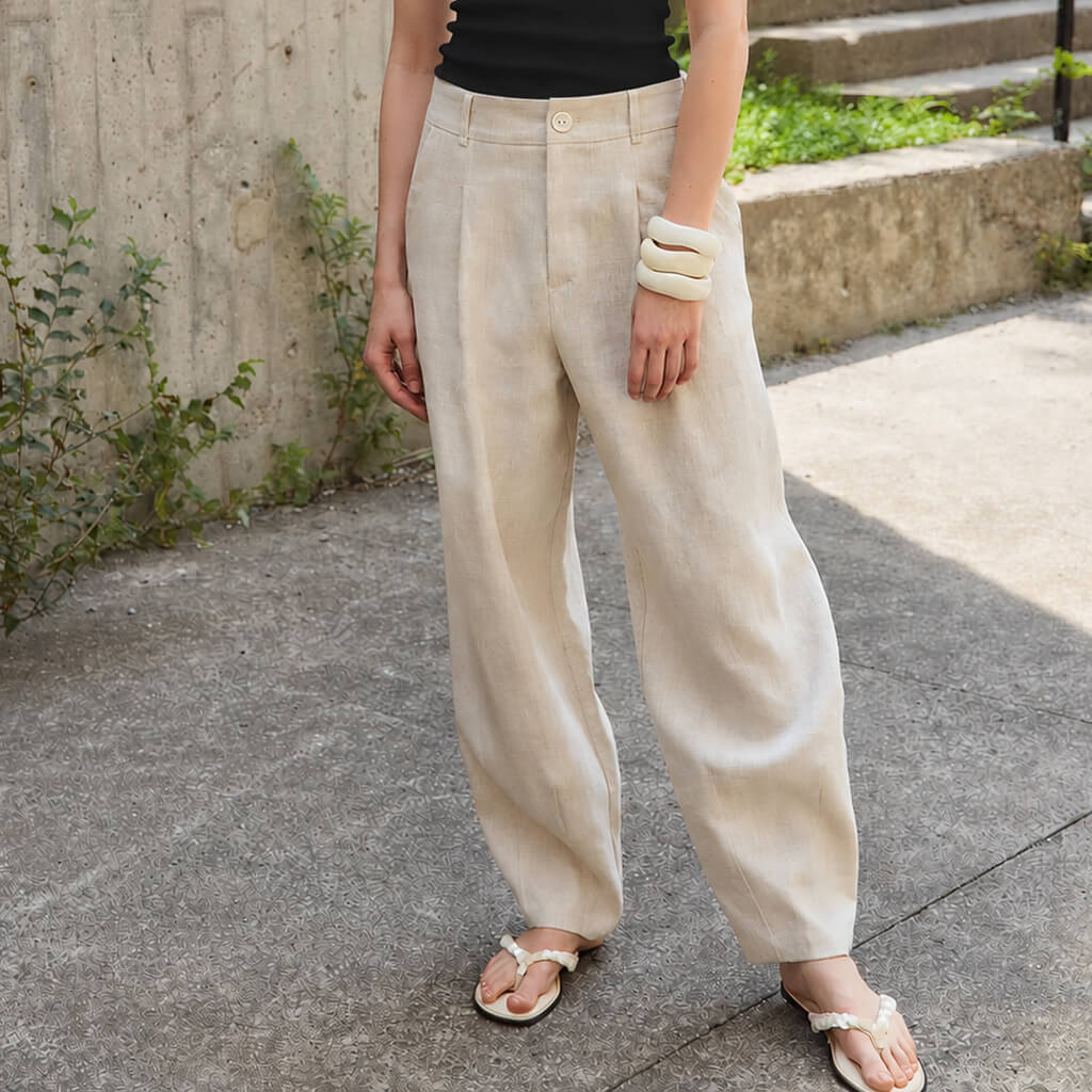 A person wearing loose, barrel-leg beige linen pants with a black top and white sandals, standing outdoors on a concrete path.