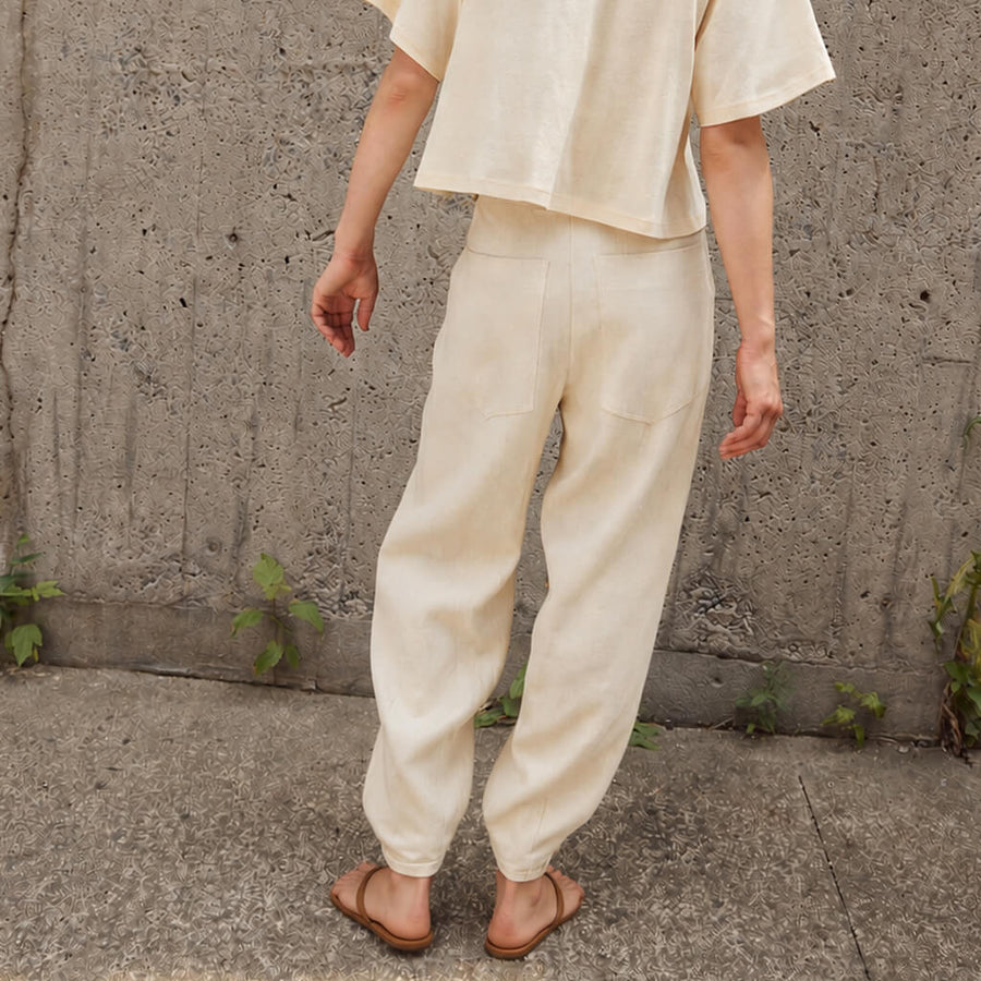 A person wearing loose, barrel-leg beige linen pants with a black top and white sandals, standing outdoors on a concrete path.