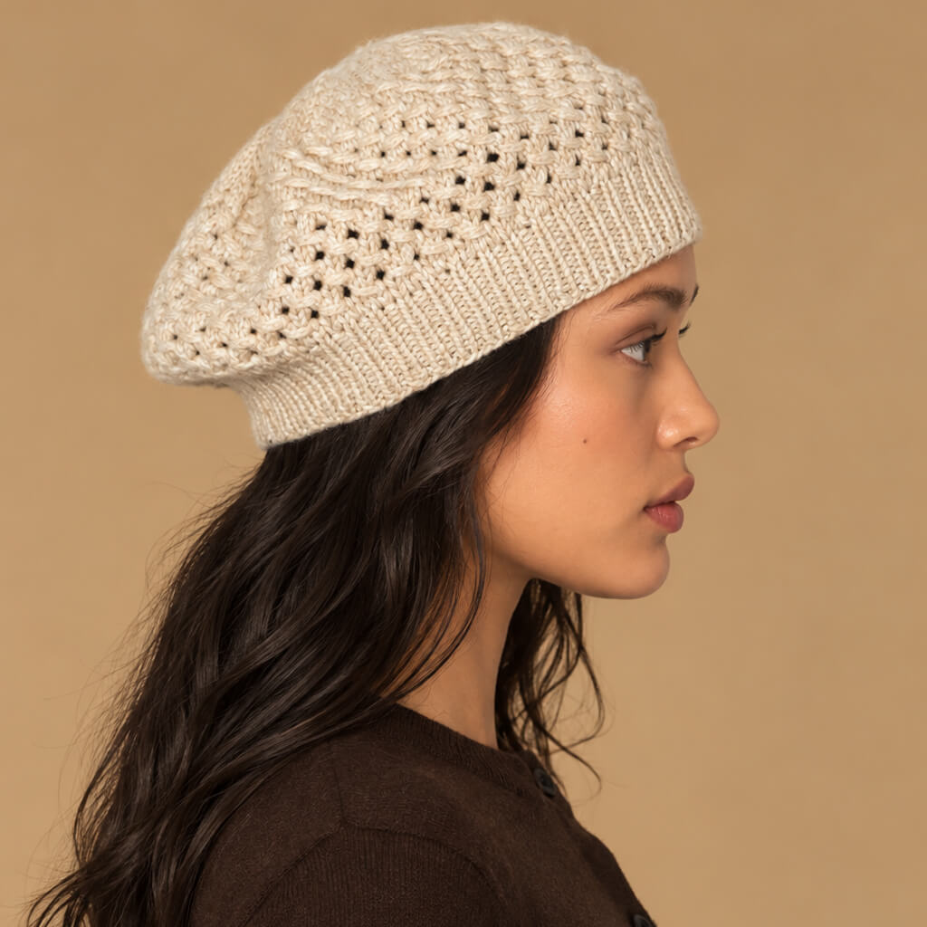 A woman in profile wearing a cream-colored, open-knit beret-style hat against a solid tan background.