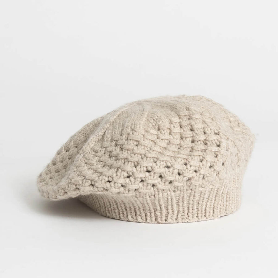 A woman in profile wearing a cream-colored, open-knit beret-style hat against a solid tan background.