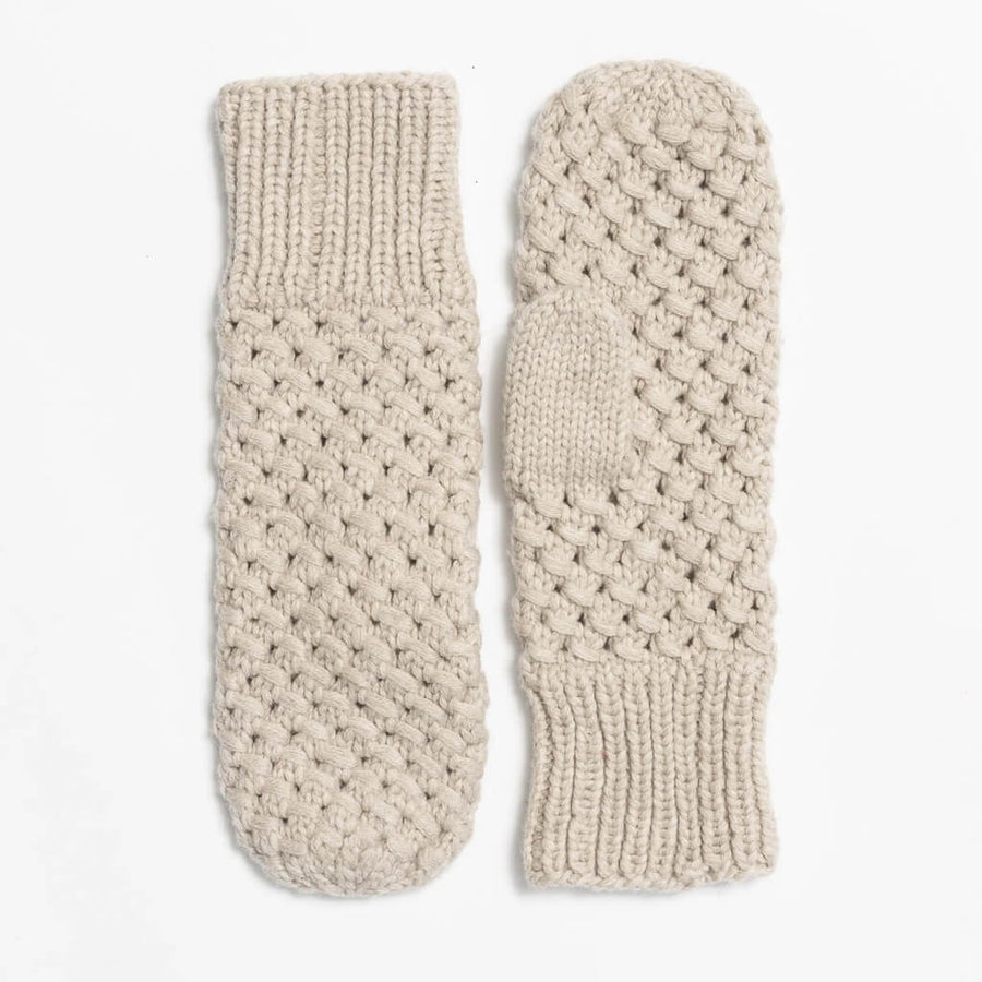 A close-up of a person wearing a cream-colored knit mitten with a textured openwork pattern, paired with white pants and a dark brown top, set against a solid tan background.