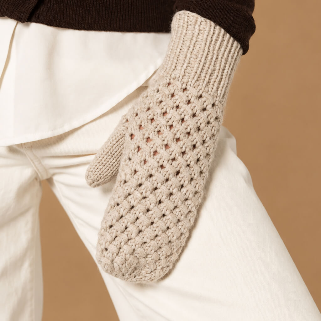 A close-up of a person wearing a cream-colored knit mitten with a textured openwork pattern, paired with white pants and a dark brown top, set against a solid tan background.
