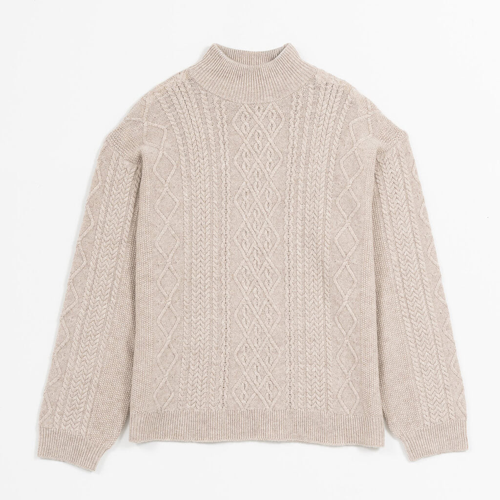 A woman wearing a beige cable-knit mock neck sweater with a diamond pattern on the chest, paired with blue jeans, set against a solid tan background.