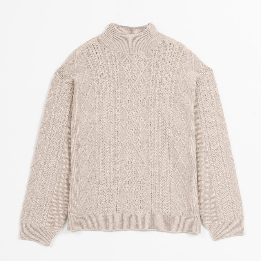 A woman wearing a beige cable-knit mock neck sweater with a diamond pattern on the chest, paired with blue jeans, set against a solid tan background.
