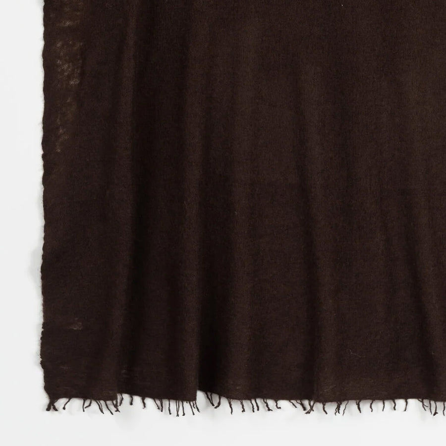 A woman wearing a dark brown fringed cashmere scarf wrapped around a cream ribbed knit sweater, posed against a solid tan background.