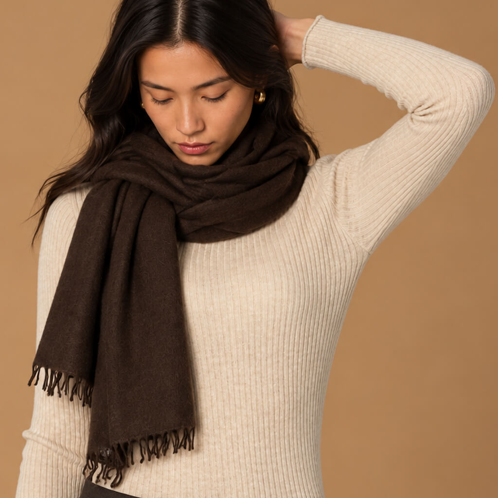 A woman wearing a dark brown fringed cashmere scarf wrapped around a cream ribbed knit sweater, posed against a solid tan background.
