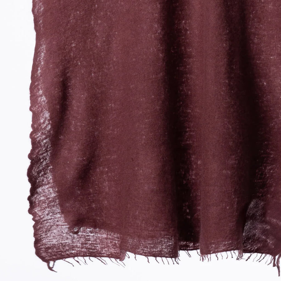 A woman wearing a dark burgundy cashmere scarf with fringed edges over a black knit sweater, set against a solid warm brown background.