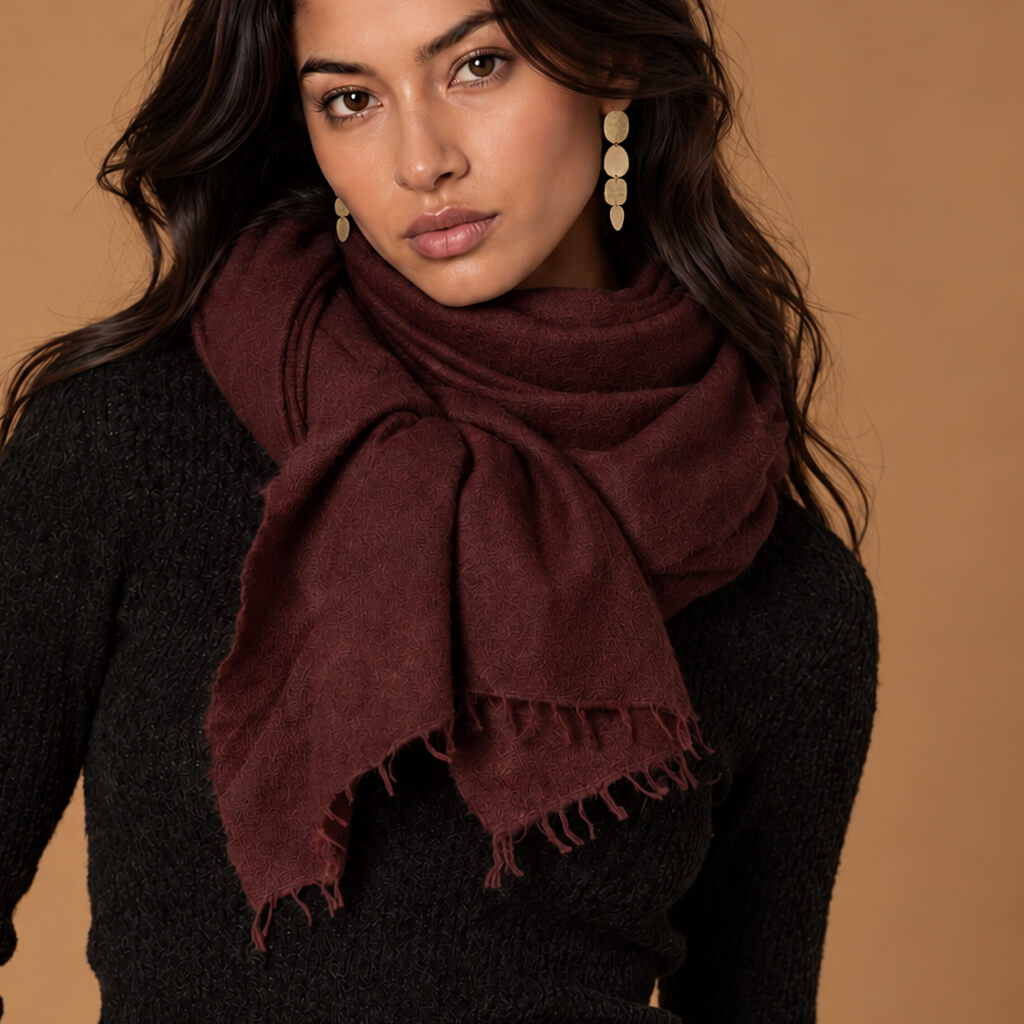 A woman wearing a dark burgundy cashmere scarf with fringed edges over a black knit sweater, set against a solid warm brown background.