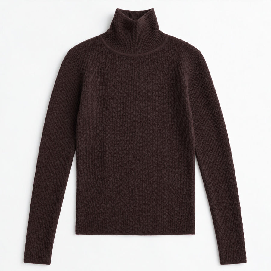 A woman with long dark hair is shown from the side in a studio setting, wearing a fitted ribbed turtleneck sweater in dark brown with light blue jeans against a solid tan background.