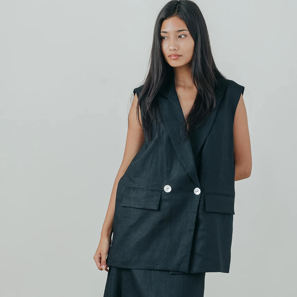A woman modeling a sleeveless black double-breasted vest with silver buttons, paired with matching dark trousers, against a light gray studio background.