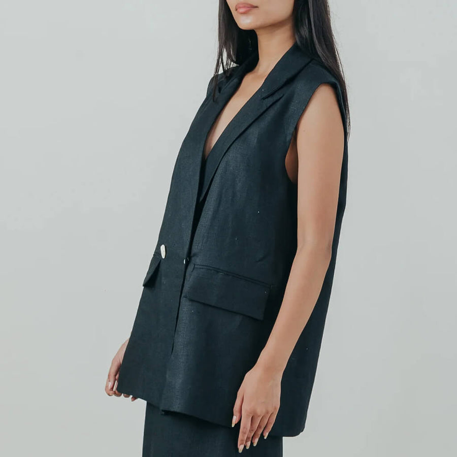 A woman modeling a sleeveless black double-breasted vest with silver buttons, paired with matching dark trousers, against a light gray studio background.