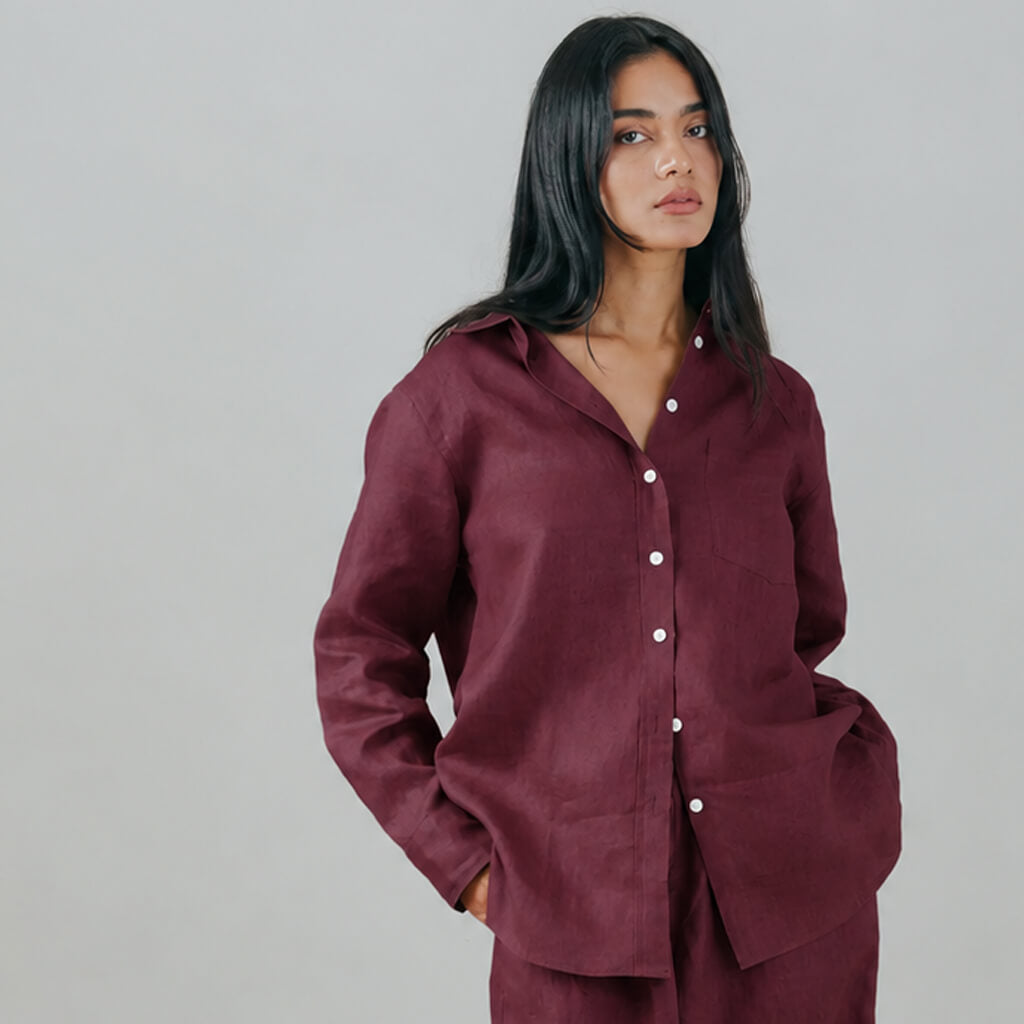 A woman modeling a loose-fitting, long-sleeve button-up shirt in a deep burgundy or maroon shade, paired with matching trousers, against a solid light gray studio background.