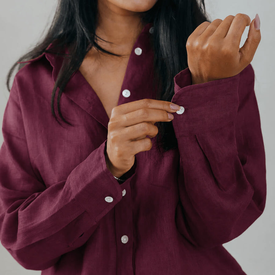 A woman modeling a loose-fitting, long-sleeve button-up shirt in a deep burgundy or maroon shade, paired with matching trousers, against a solid light gray studio background.