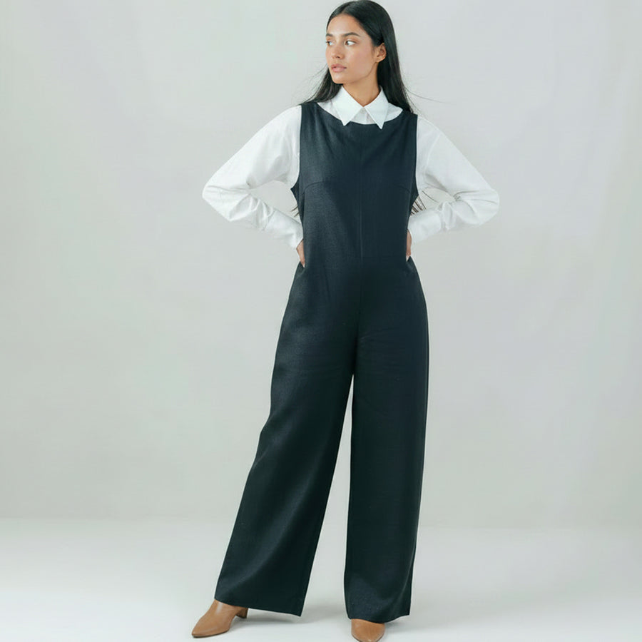 A woman modeling a black sleeveless jumpsuit with wide legs, standing against a neutral light gray studio background.
