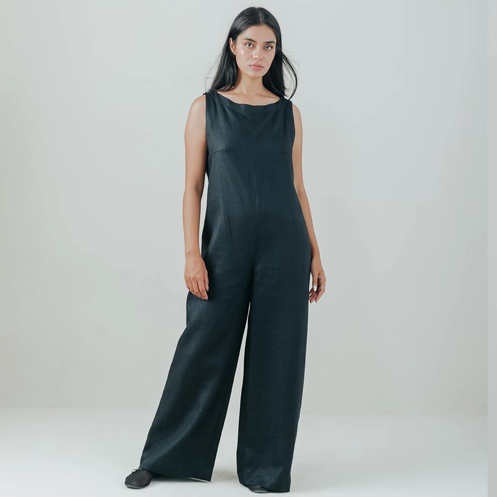 A woman modeling a black sleeveless jumpsuit with wide legs, standing against a neutral light gray studio background.