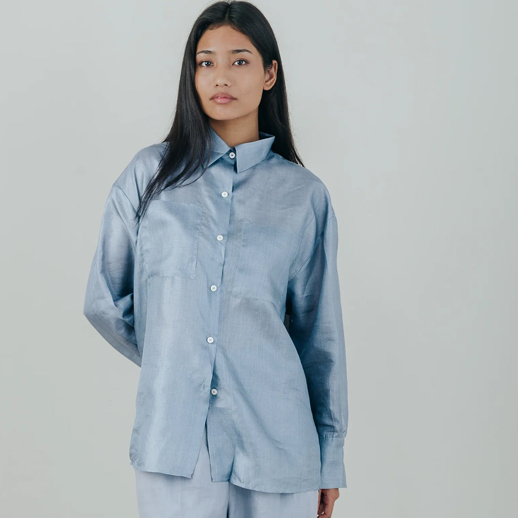 A woman modeling a light blue long-sleeve button-up shirt with a high collar, photographed against a plain light gray studio background.