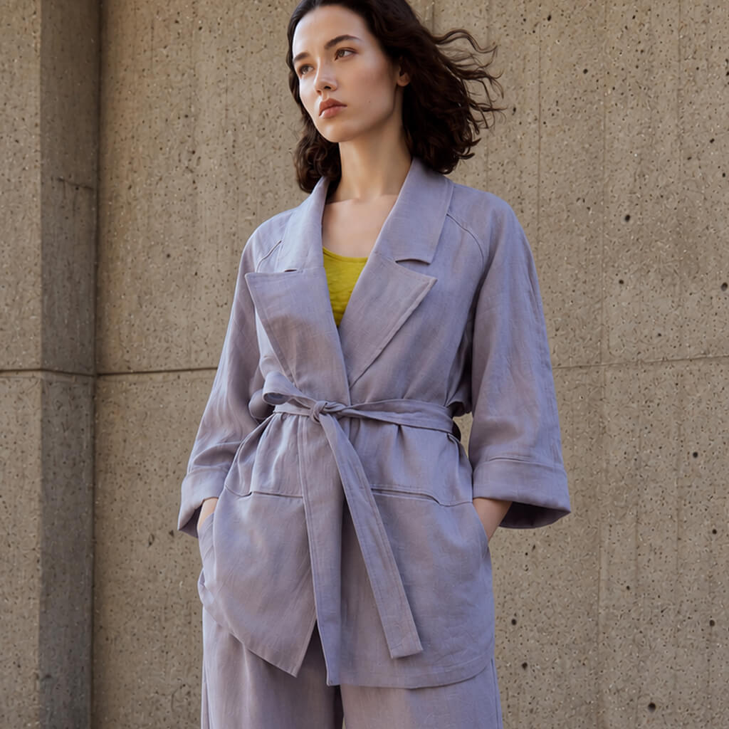 A woman wearing a light lavender-gray belted jacket and matching pants, standing against a textured concrete wall.