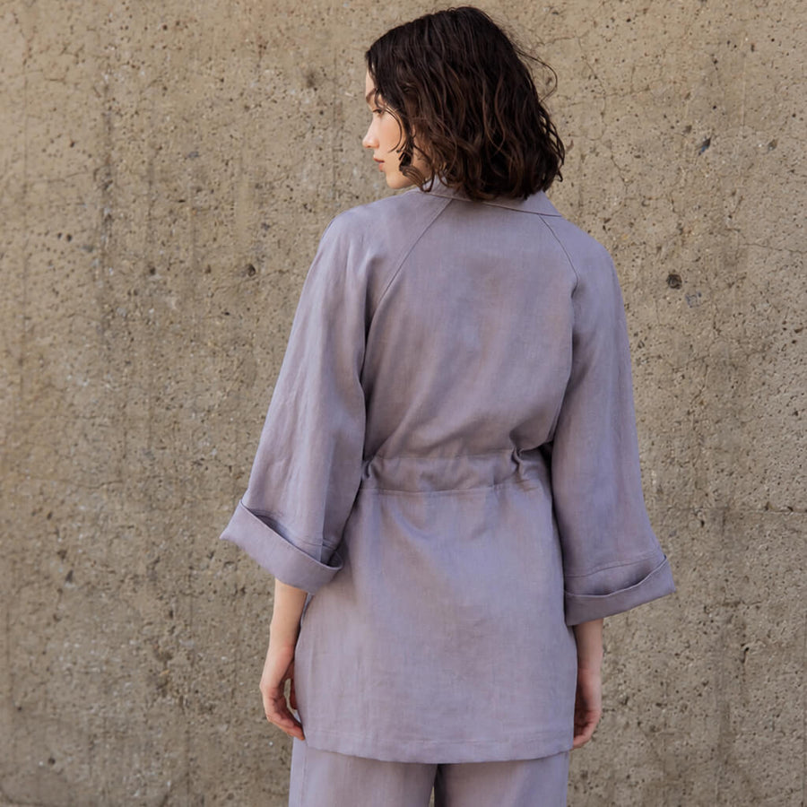 A woman wearing a light lavender-gray belted jacket and matching pants, standing against a textured concrete wall.