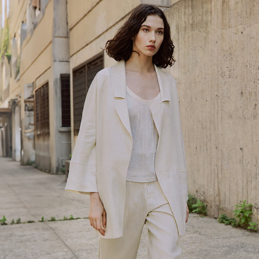 A woman wearing a cream-colored, oversized belted coat and matching trousers, photographed outdoors against a concrete building wall.