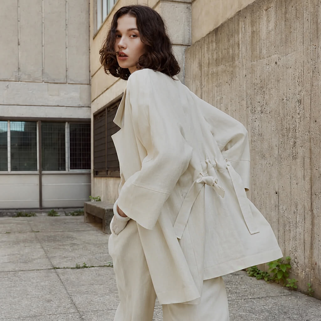 A woman wearing a cream-colored, oversized belted coat and matching trousers, photographed outdoors against a concrete building wall.